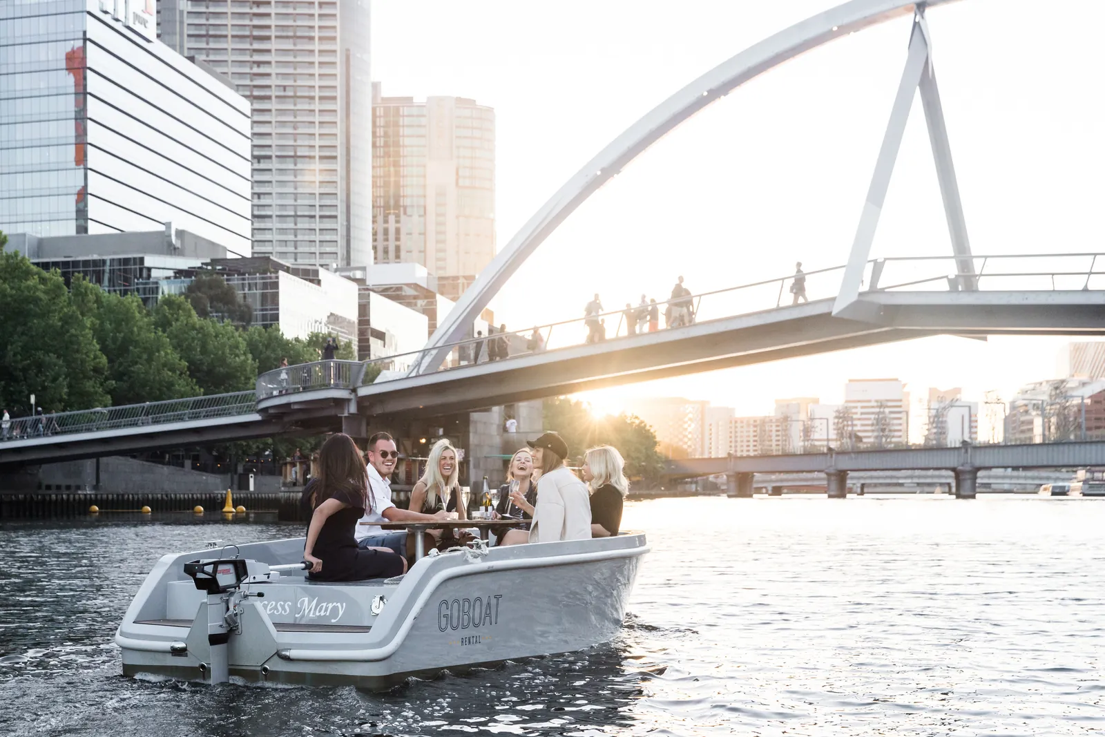 A GoBoat electric picnic boat on the Yarra below a footbridge, Melbourne's towers behind