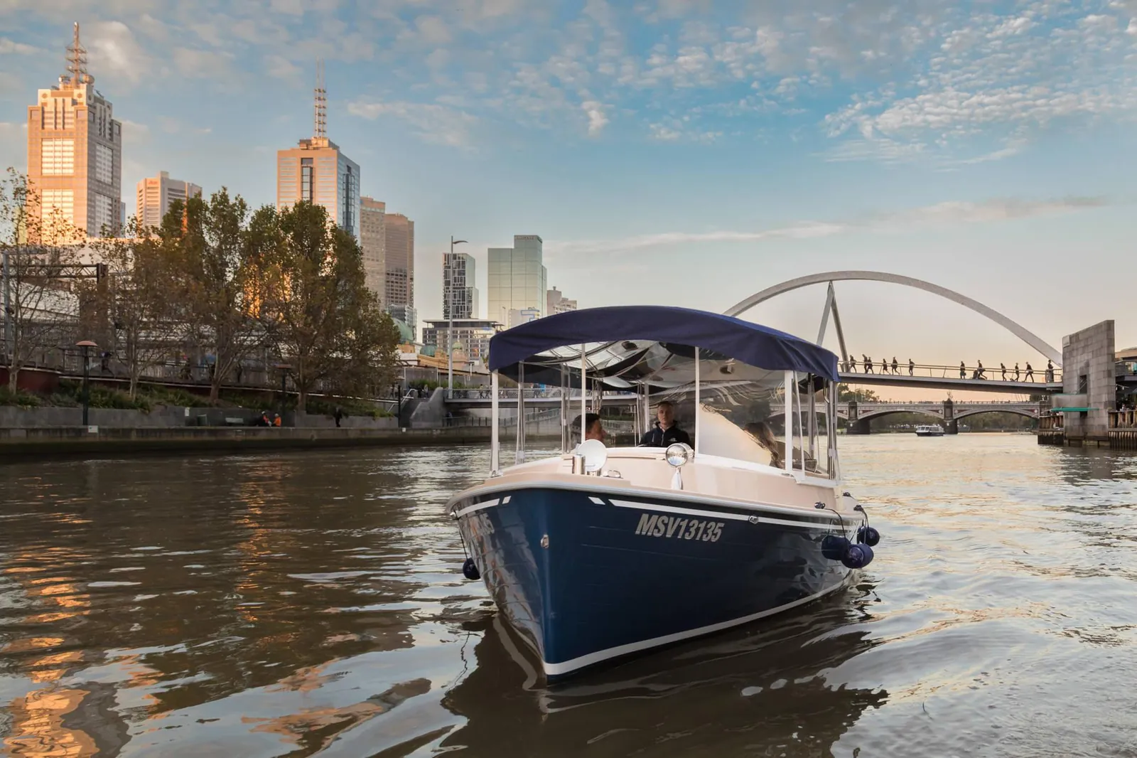A small electric charter boat on the Yarra with riverside towers behind